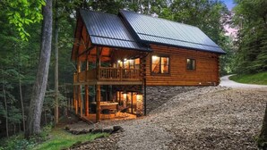 A log cabin with a balcony, a stone foundation, and a gravel driveway.