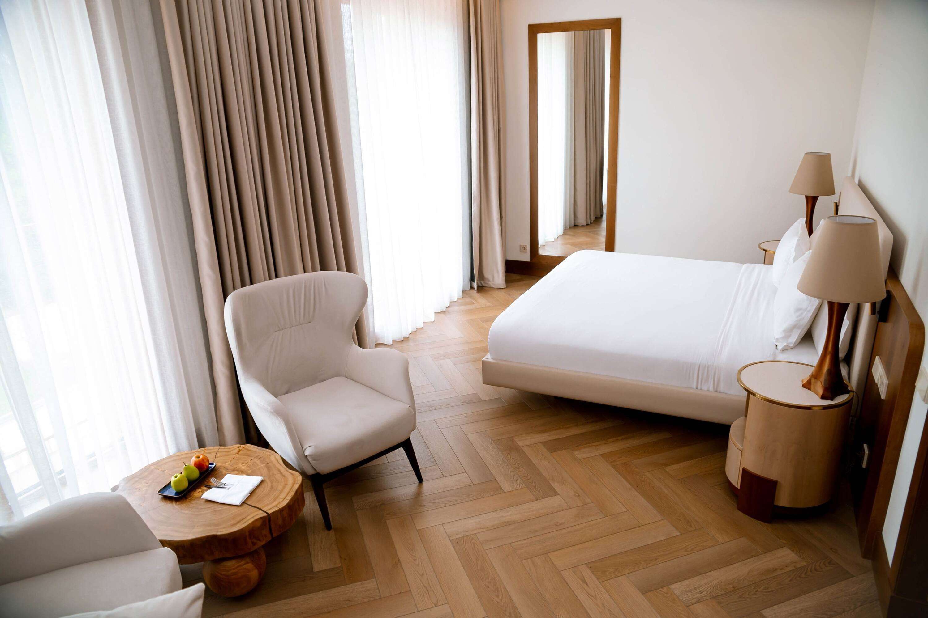 A hotel room with a bed, a chair, a round wooden table with fruit, and a lamp.