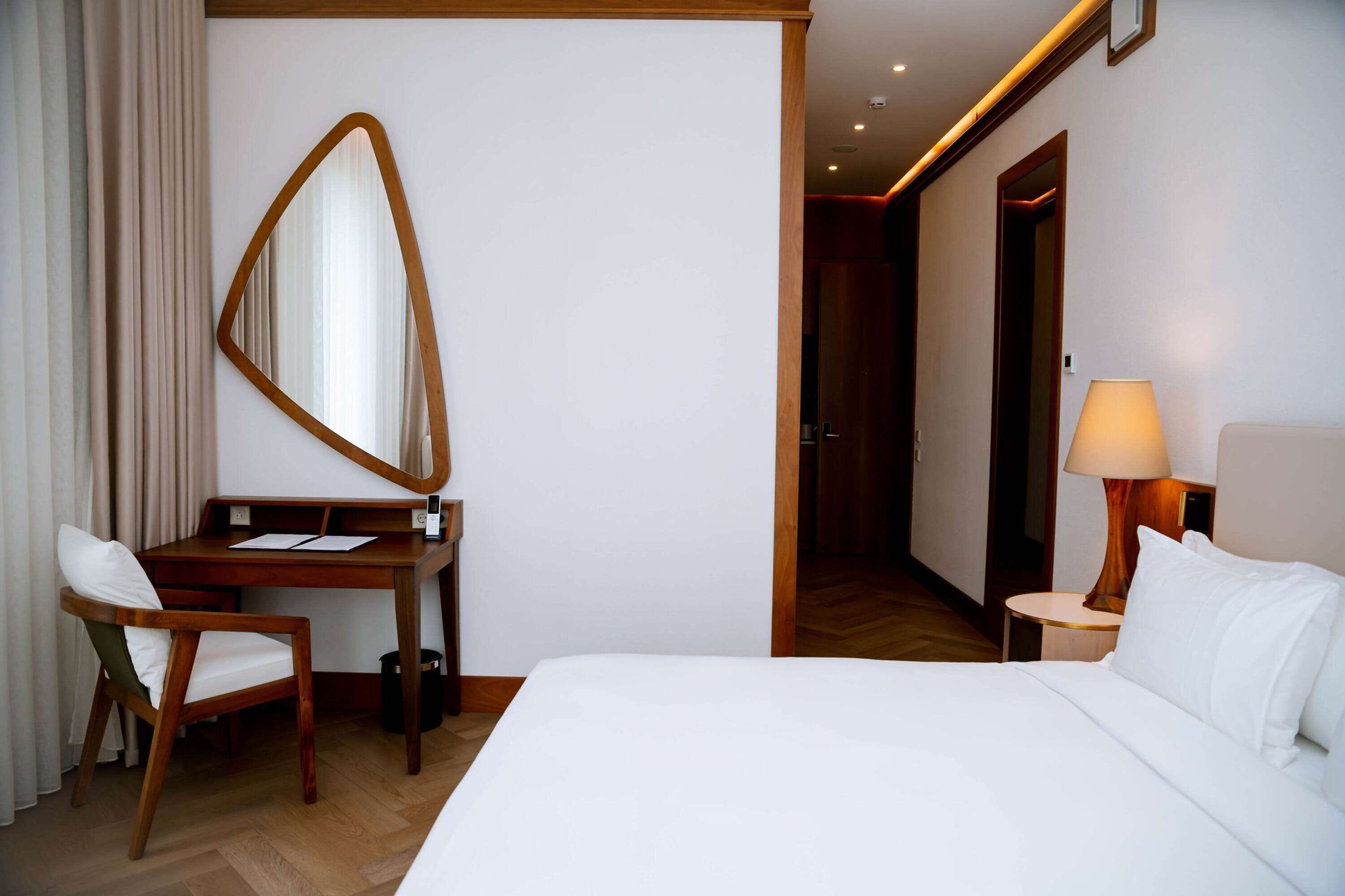Habitación de hotel con cama, escritorio, silla, espejo, lámpara y puerta que da a otra habitación.