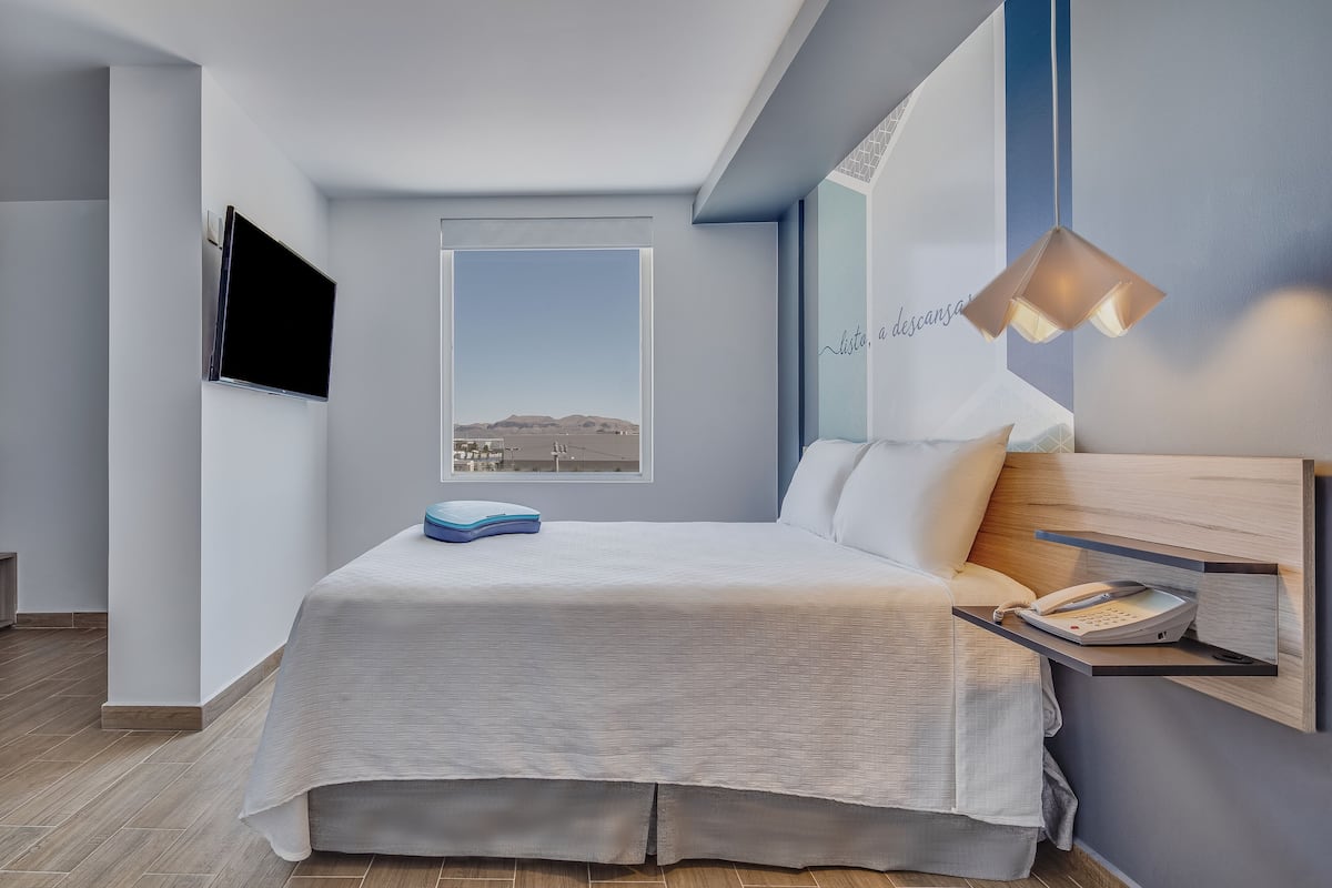 A modern bedroom with a bed, a mounted TV, a bedside telephone, and a view of the landscape through a window.