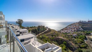 Vista costera con cielo despejado, un edificio con balcón de vidrio que da al mar y una colina con edificaciones residenciales.