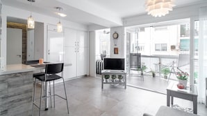 A modern kitchen with white cabinets, a dining area with a black chair, and a living room with a sofa and a TV.