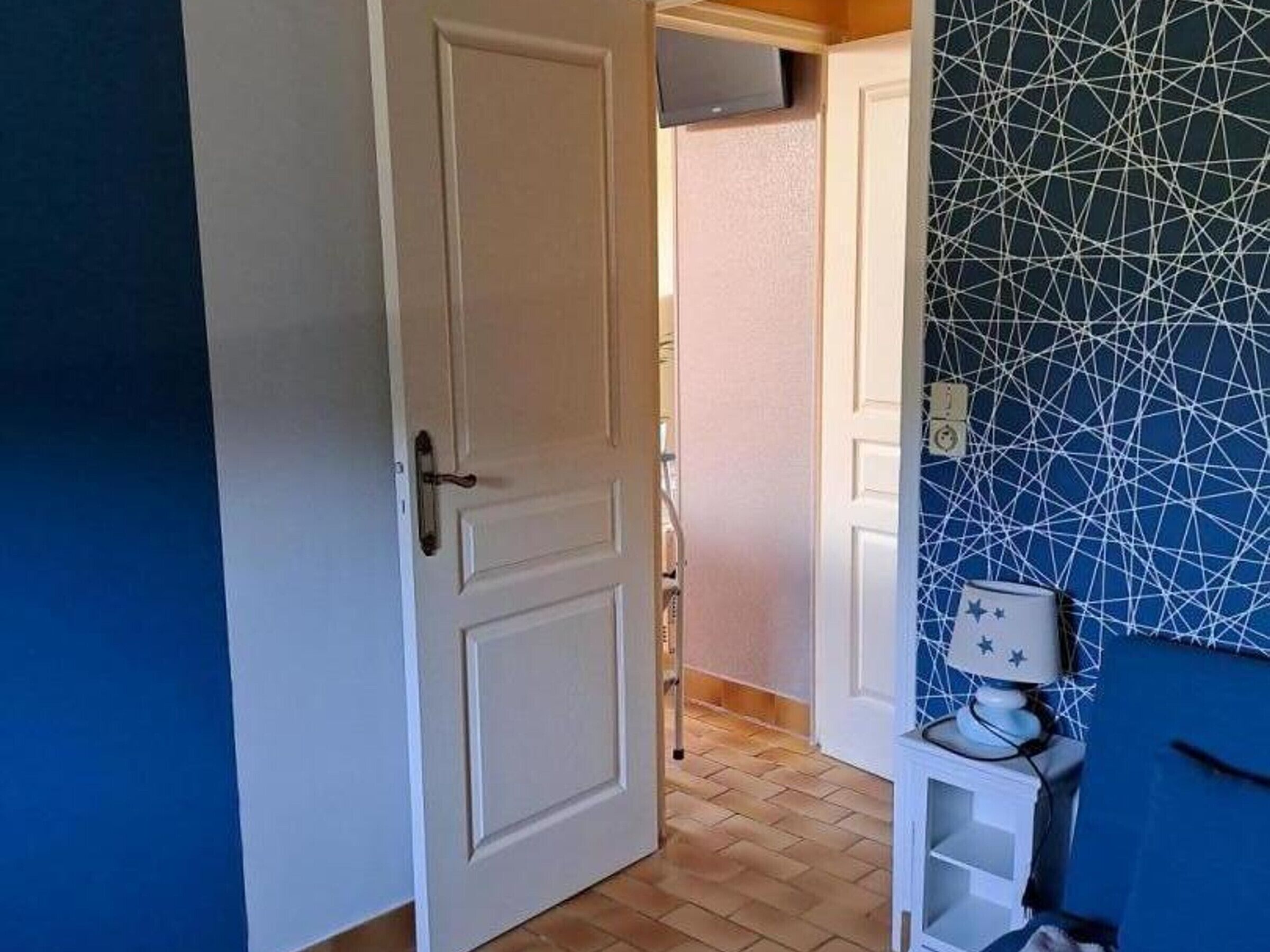 Een kamer met een witte deur, een klein wit kastje met een lamp en een blauwe, patroonrijke wand.
