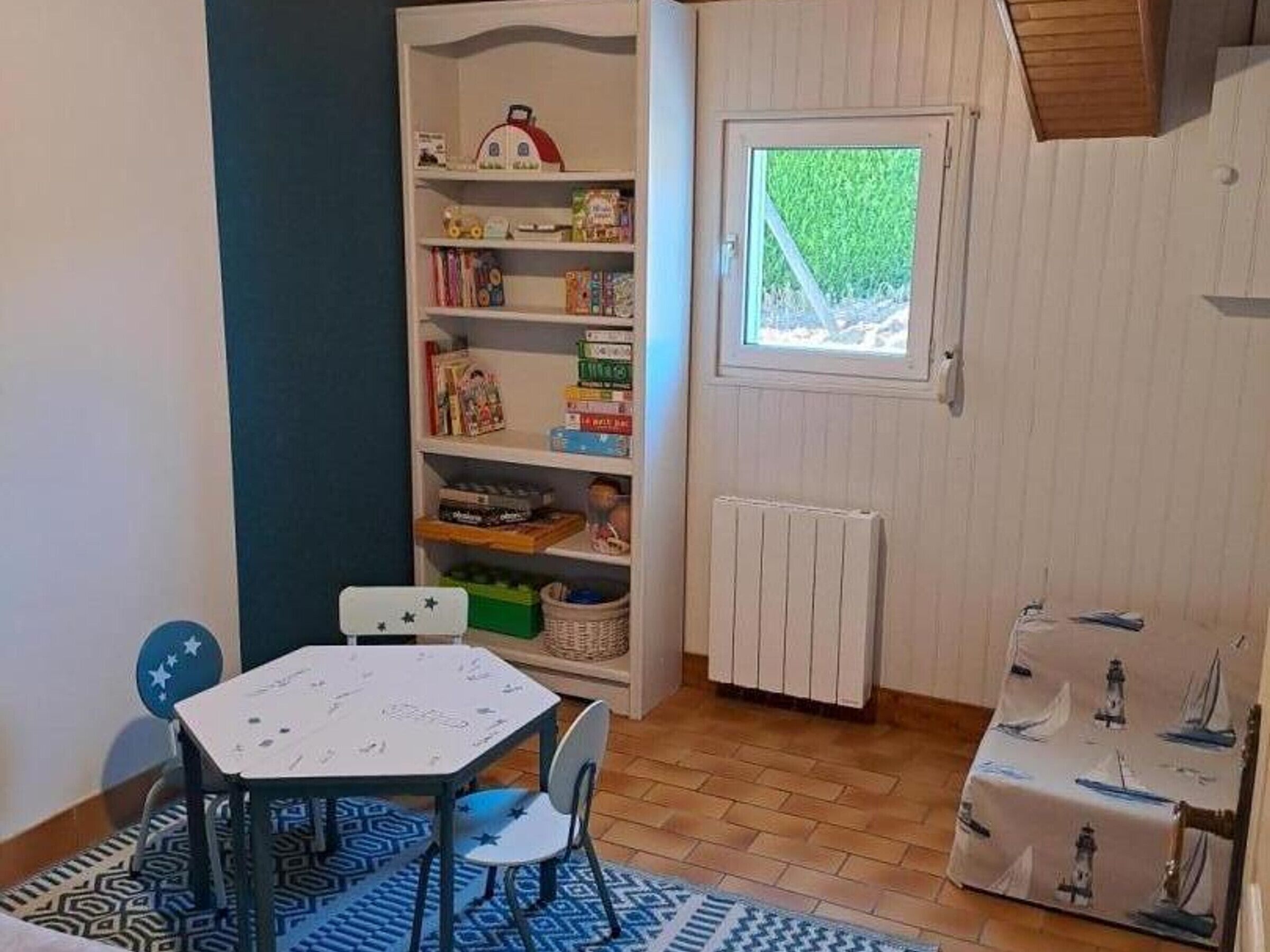 A room with a white radiator, a window with a view of greenery, a white table with chairs, and a bookshelf filled with various items.