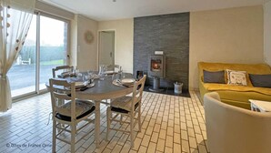 A dining area with a round table and chairs, a sofa, a fireplace, and a view through a large window. - Charming granite house with enclosed garden, all inclusive, near Domfront (Saint-Mars-d'Égrenne)