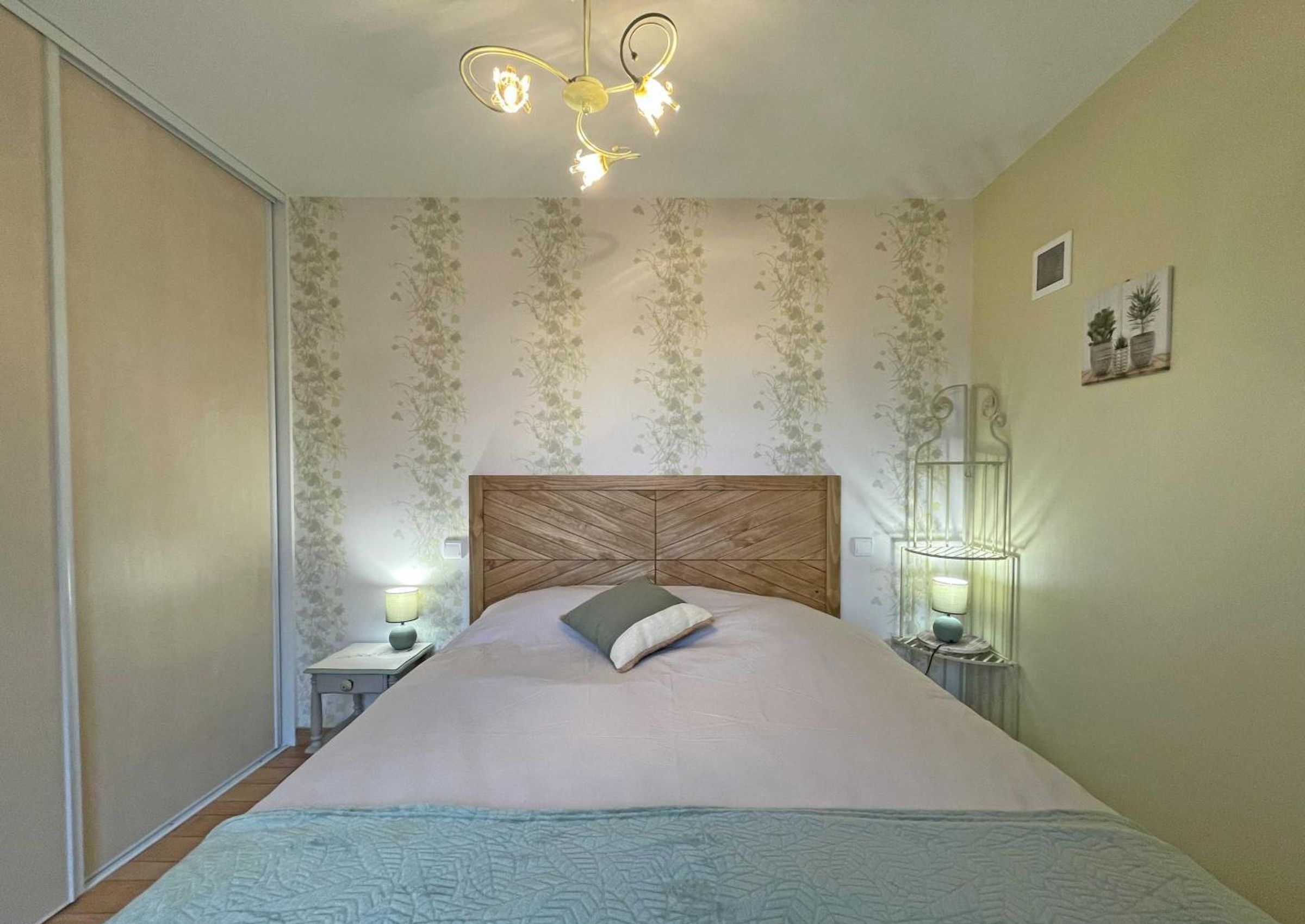 A bedroom with a wooden headboard, floral wallpaper, two bedside lamps, and a bed with a blue quilt.
