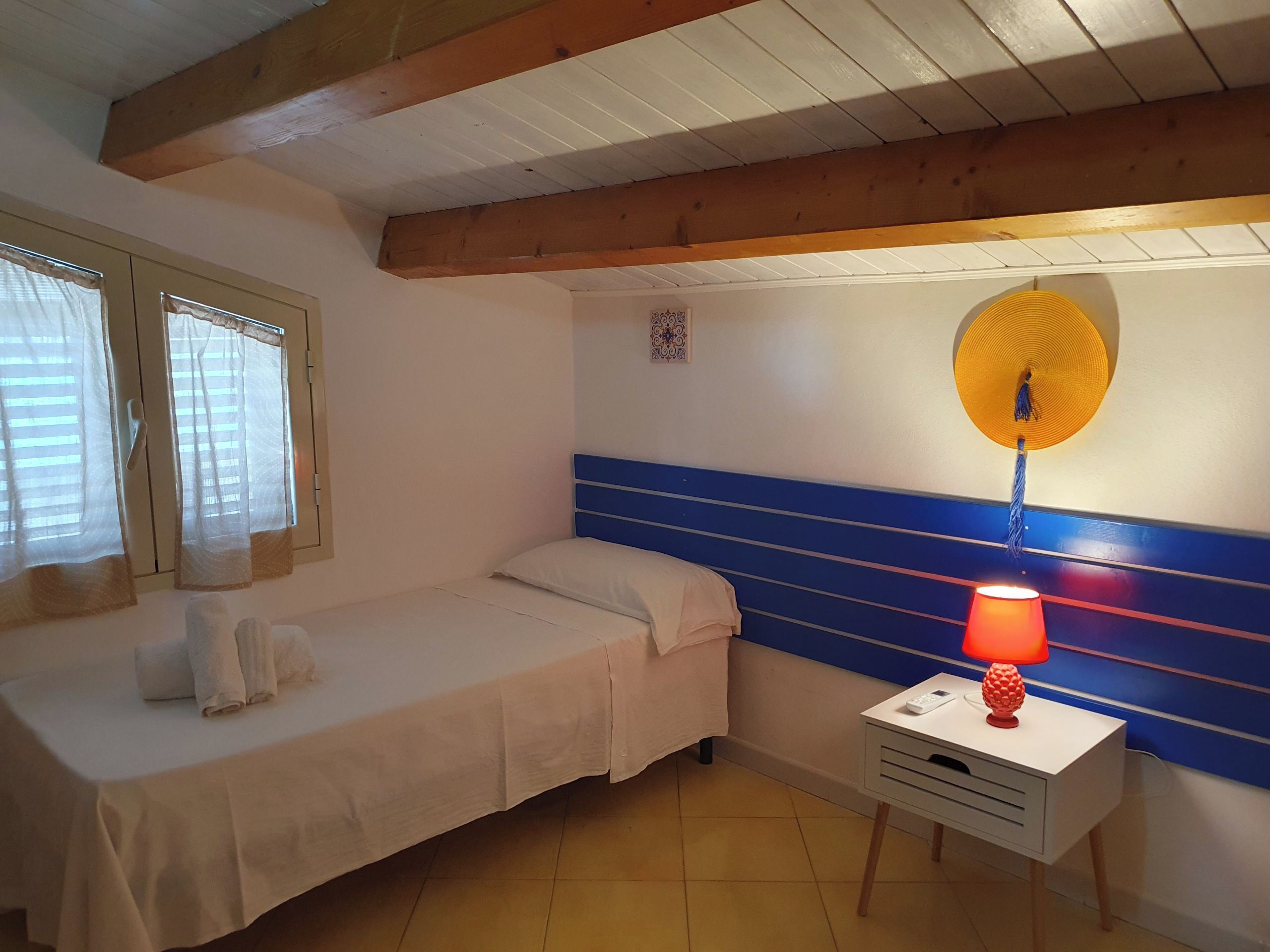Una camera piccola con un letto singolo, soffitto in legno, comodino con lampada e finestra con tende.