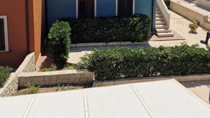 A two-story building with a tiled roof, a small garden area with bushes, and a staircase leading to the upper floor. - A6 third row foreground (Marzamemi)