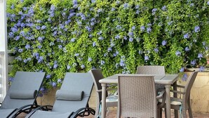 A patio area with a table and chairs, a brick floor, and a wall covered with green foliage and blue flowers. - Apartment D2 sea view first floor (Marzamemi)