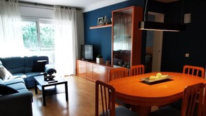 Ein Wohnzimmer mit blauem Sofa, Couchtisch, Esstisch mit Stühlen, Fernseher auf einer Kommode und einem Fenster mit Vorhängen.