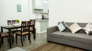 Een moderne woonkamer met een grijze bank, een houten eettafel en stoelen. Er is een keuken zichtbaar op de achtergrond met witte kasten en een aanrecht.