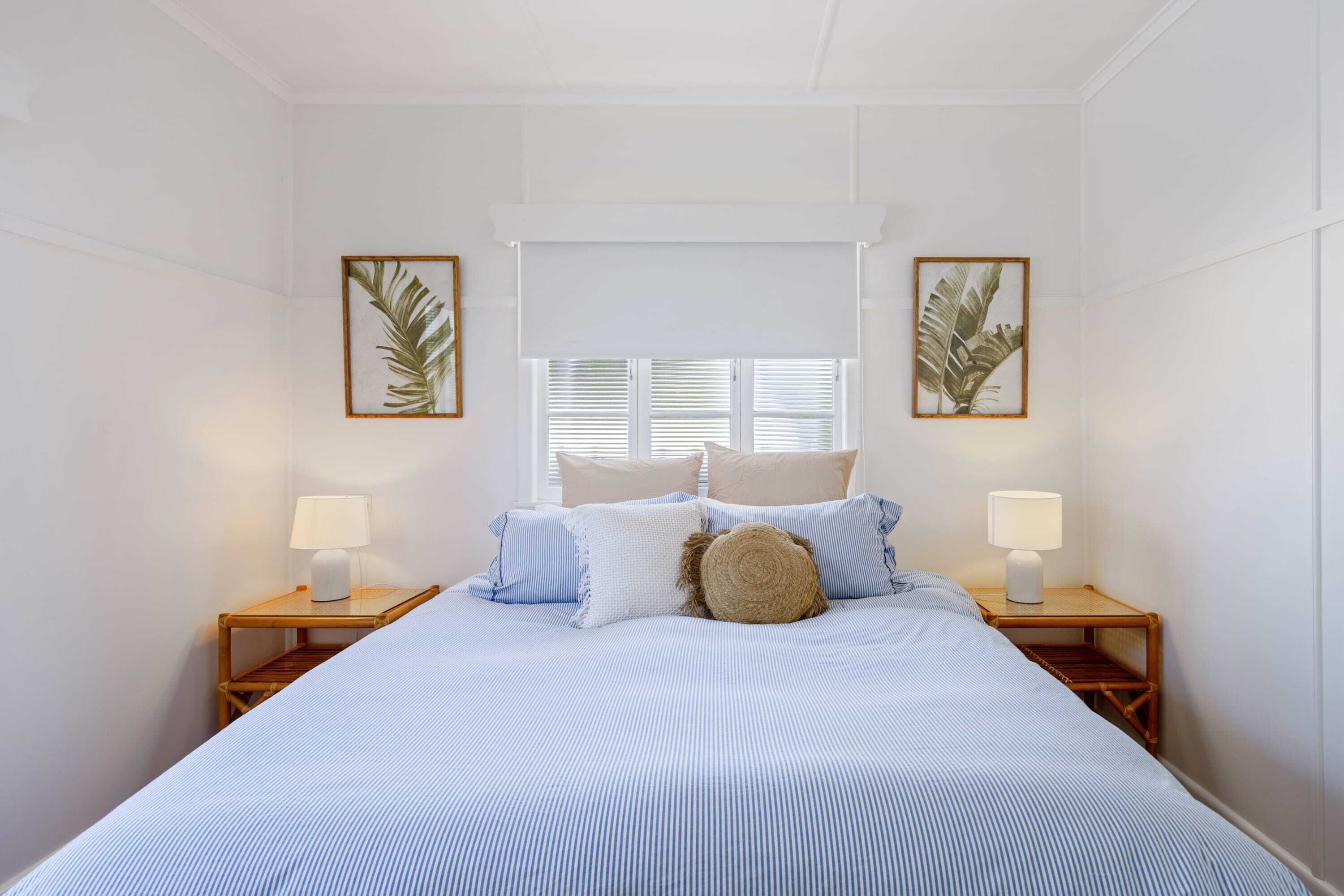 A bedroom with a bed, two bedside tables with lamps, a window with blinds, and framed pictures on the wall.