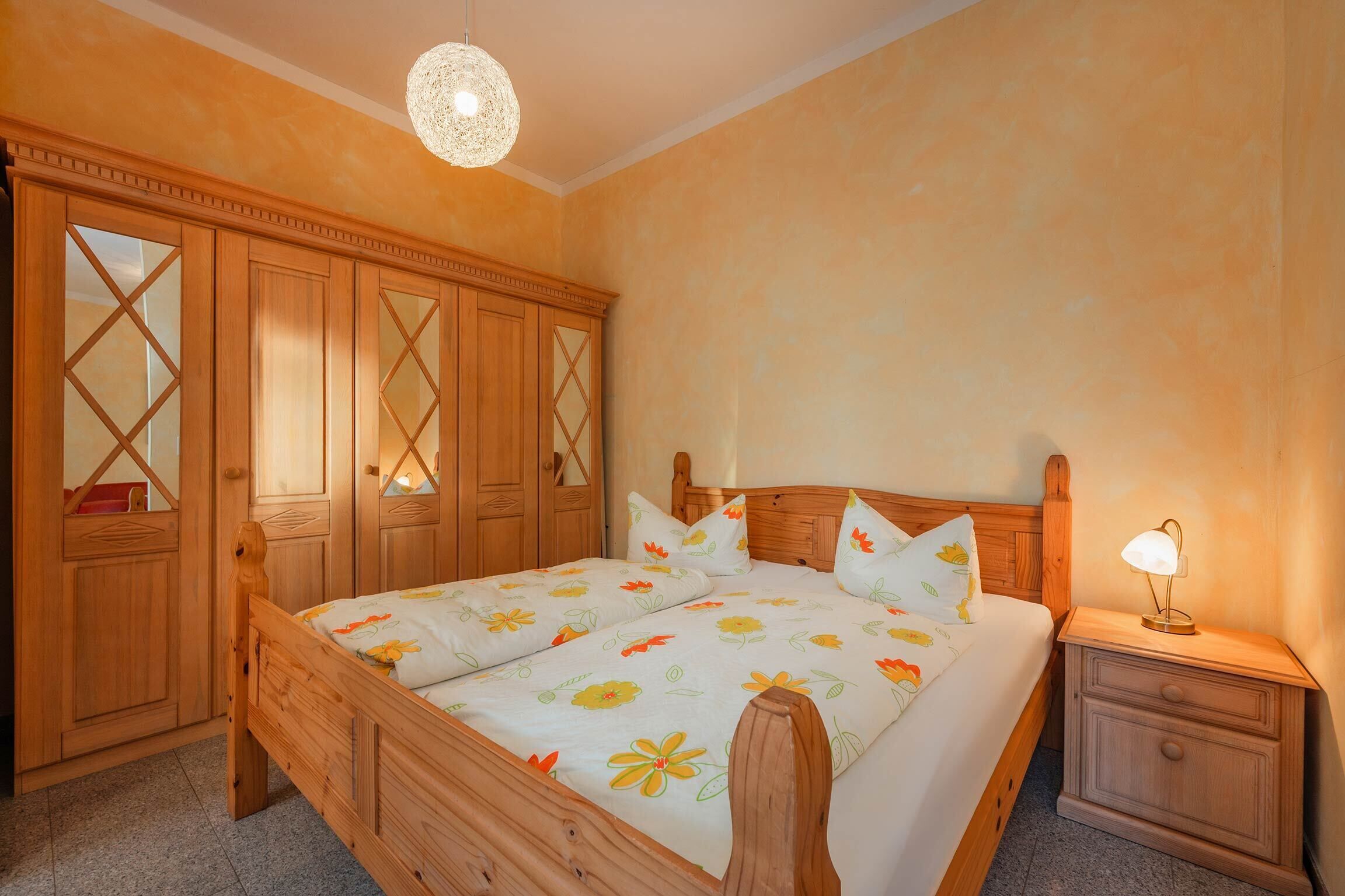 Ein Bett mit Holzbettgestell, mit Blumenmuster bezogene Bettwäsche, ein Nachttisch mit Lampe und ein Schrank mit Spiegelüren.