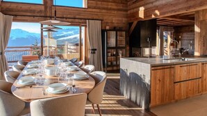 A wooden cabin with a dining area, kitchen, and large windows overlooking a mountainous landscape. - Luxury 10 person, 5 bedroom chalet with hot tub, sauna and steam room. (Landry)