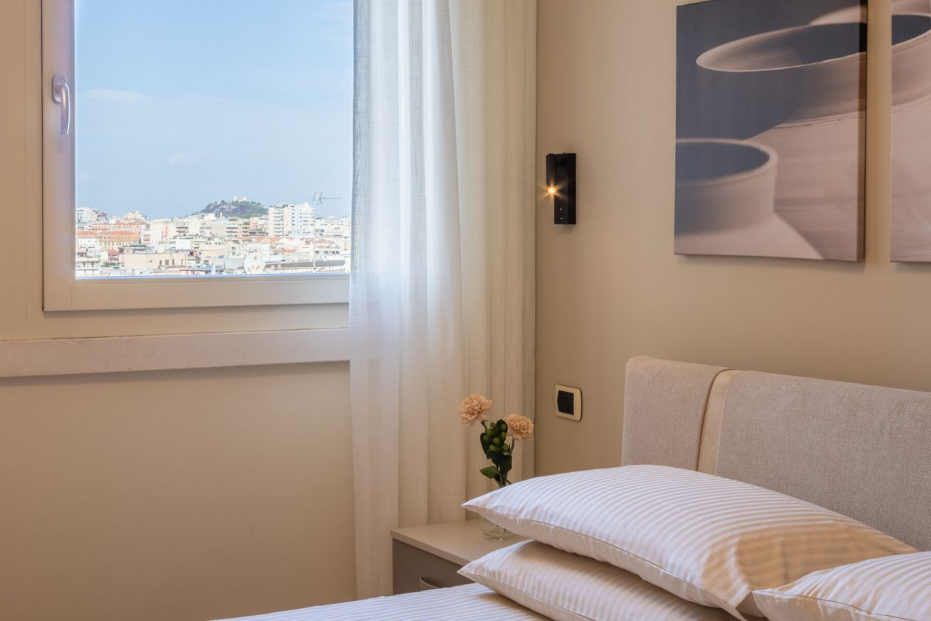 Um quarto com cama, mesa de cabeceira, um vaso com flores, janela com vista para a cidade e luminária na parede.