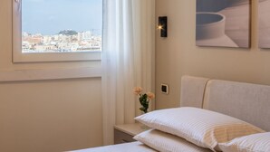 Una camera da letto con un letto, un comodino, un vaso con fiori, una finestra vista sulla città e un lampadario a parete.