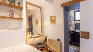 Une chambre à coucher comprenant un lit, une chaise, un grand miroir et un radiateur. On y trouve également des étagères ornées d’objets décoratifs et une porte donnant sur une salle de bain dont le sol est recouvert de carreaux à motifs.