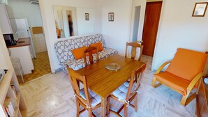 Un comedor con una mesa y sillas de madera, un sofá con cojines naranja, y una cocina al fondo.