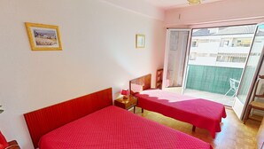 Habitación de hotel con dos camas, suelo de madera, puerta corredera de vidrio a un balcón y una mesita de noche con lámpara.