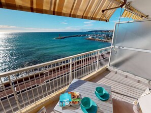 Een balkon met uitzicht op zee, een tafel met een boek en twee stoelen, en een gestreept zonnescherm.