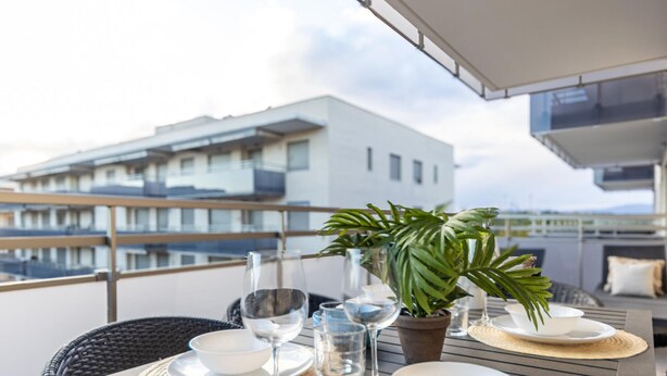 Un tavolo da pranzo apparecchiato con piatti, ciotole e bicchieri da vino su un balcone con vista su un edificio.