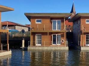 A wooden houseboat with a balcony, a dock, and a church steeple in the background.