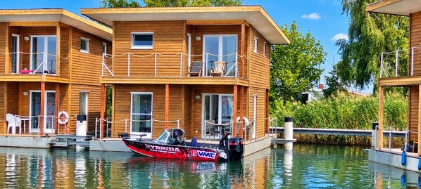A wooden houseboat with a dock, a boat named 'HYBRIDA' docked at the front, and a clear sky.