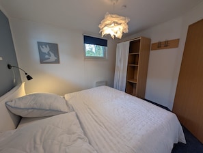 Ett sovrum med en säng, en sänglampa, ett fönster, en dörr och en hylla som är monterad på väggen.
