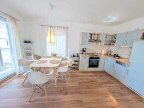 Ett modernt kök med ljusblå skåp, en matplats med ett vitt bord och stolar samt ett fönster med gardiner.