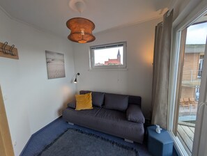 Ett modernt vardagsrum med en grå soffa, en gul kudde, en flätad lampa, en inramad tavla och ett fönster med utsikt över byggnader.