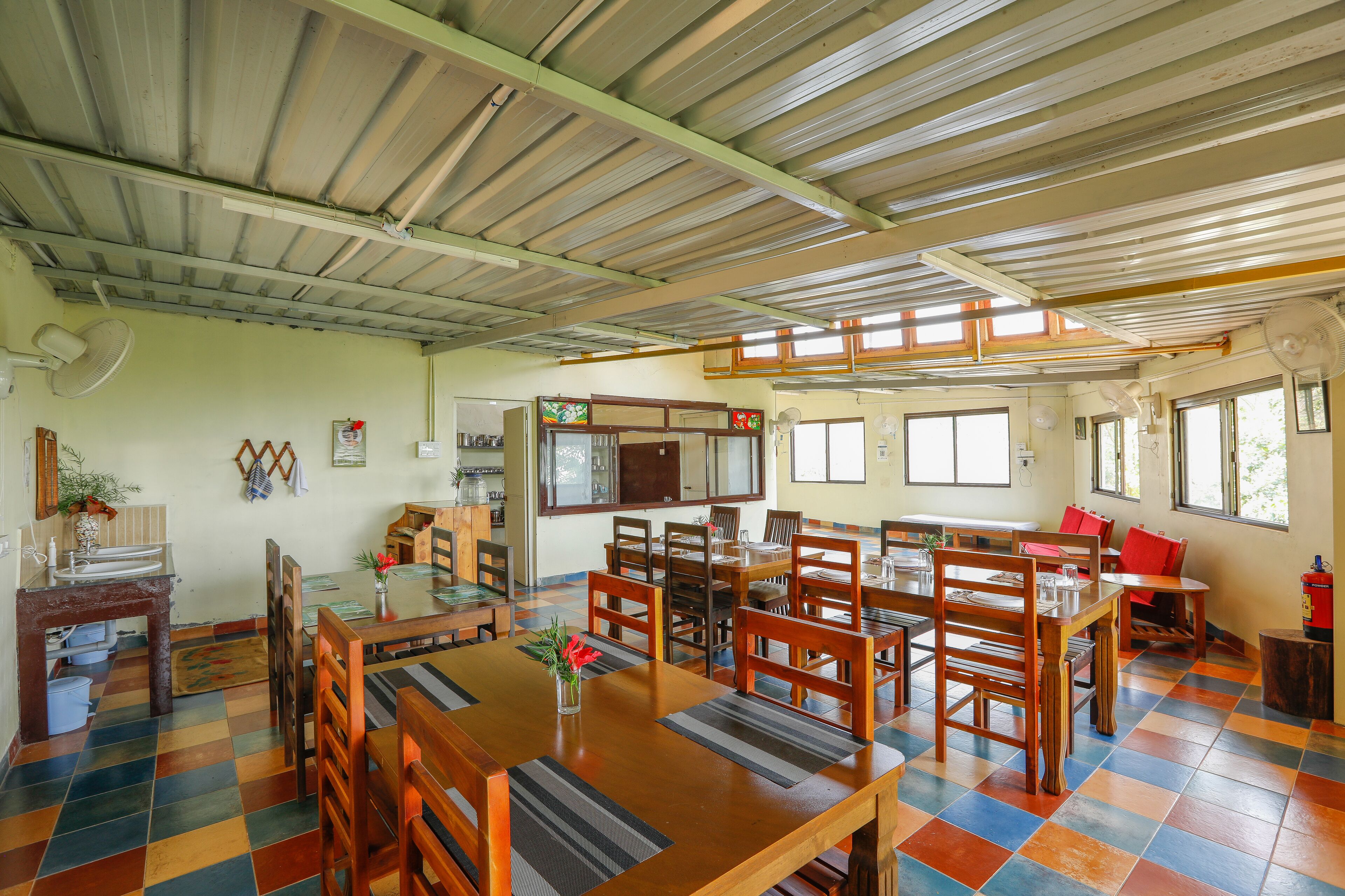 A spacious dining area with wooden tables and chairs, a kitchen in the background, and a colorful tiled floor.