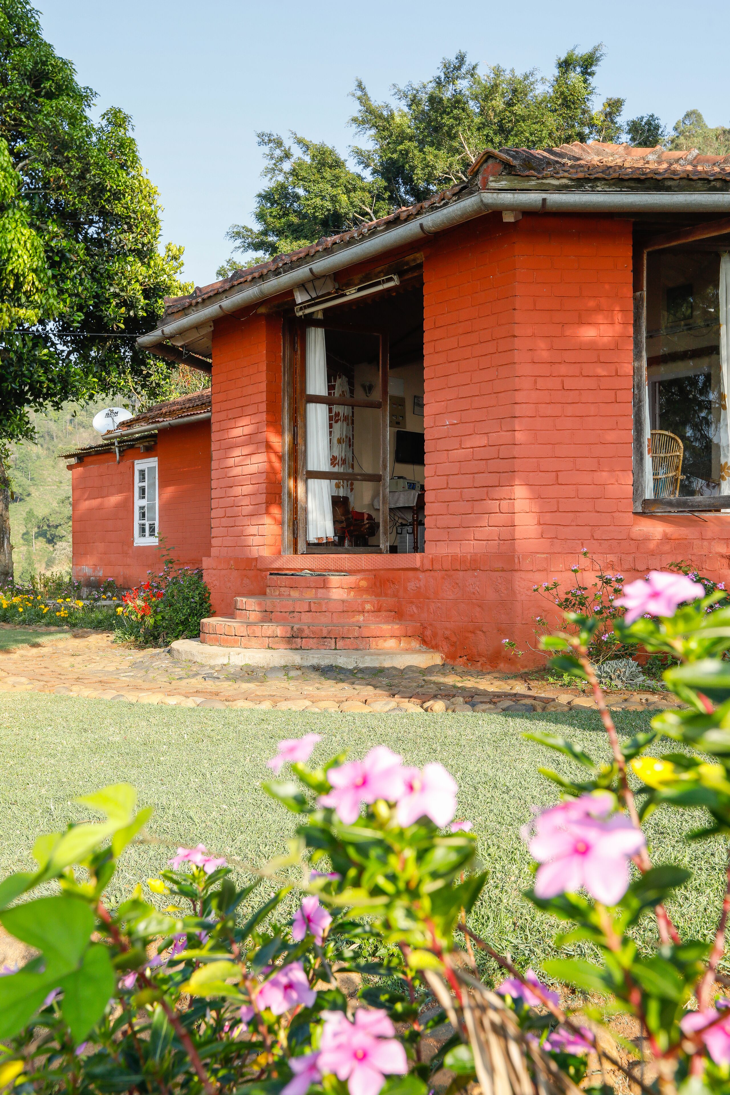 Une maison en briques rouges avec un porche, entourée d’un jardin où poussent des fleurs roses.