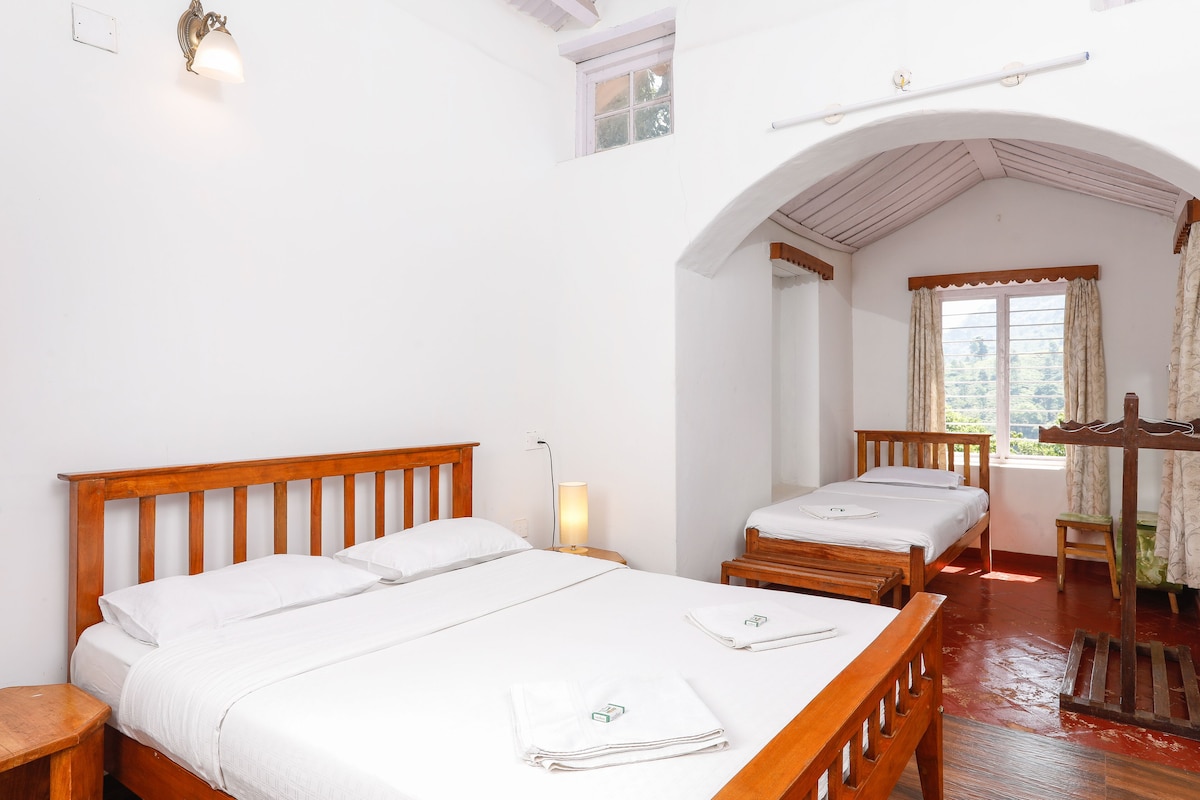 Habitación de hotel con una cama grande, una cama más pequeña, un banco de madera, una ventana con cortinas y una pequeña mesa.