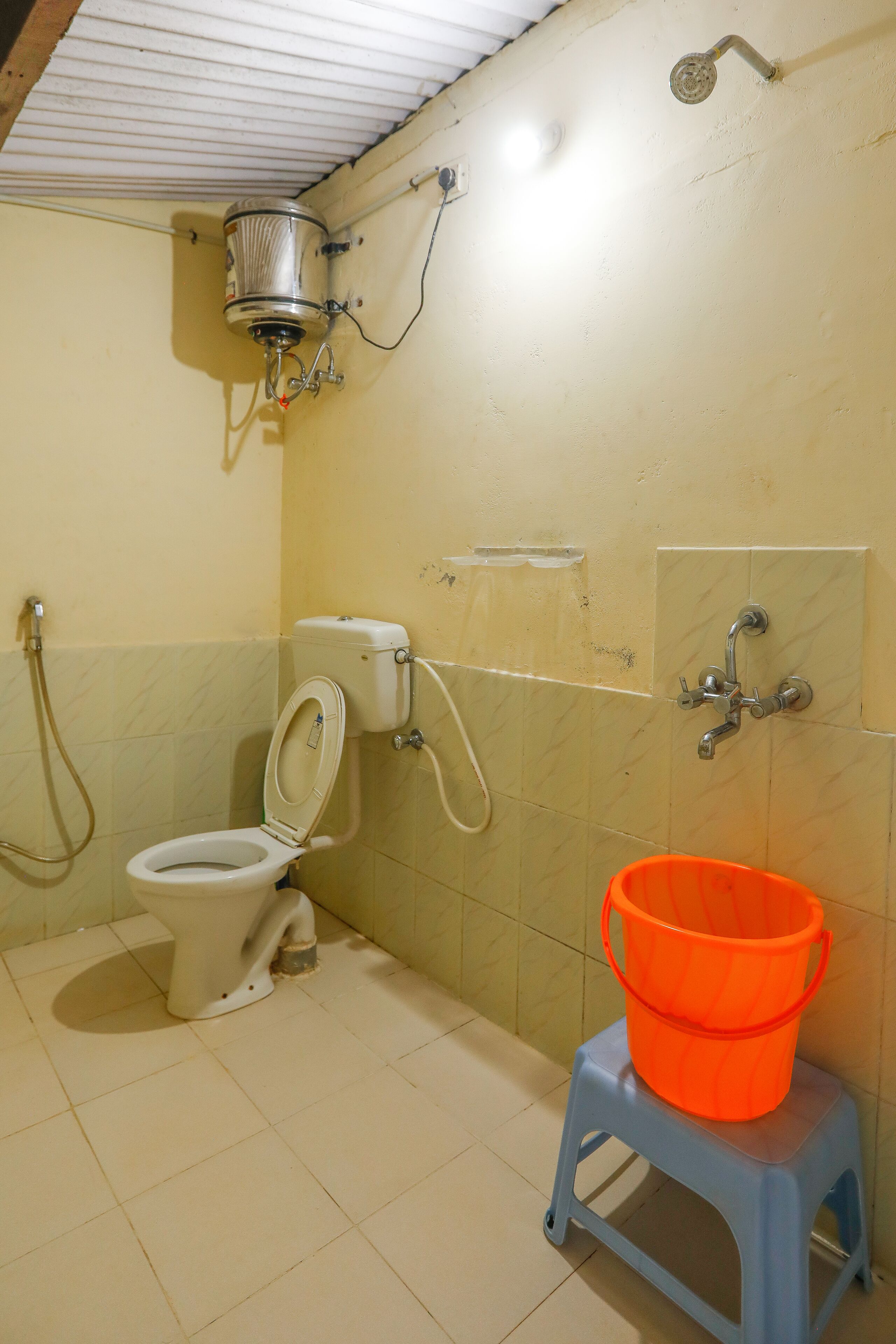Une salle de bain avec des toilettes, une douche et un petit seau orange posé sur un tabouret.