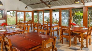 Un comedor con mesas y sillas de madera, ventanas amplias con vistas a zonas verdes y lámparas colgantes.