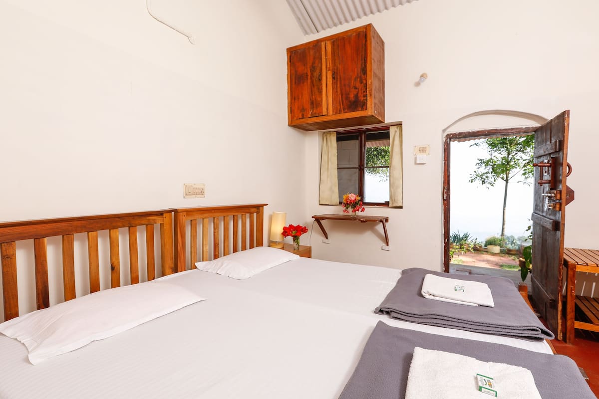 A bedroom with two beds, a wooden headboard, a small bedside table with a lamp and flowers, and a window with a view of greenery.