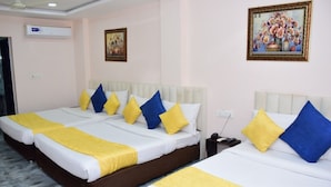Une chambre d’hôtel avec deux lits, chacun agrémenté de draps blancs et d’oreillers jaunes et bleus. On y trouve un ventilateur de plafond, un appareil de climatisation et deux œuvres d’art encadrées accrochées aux murs.