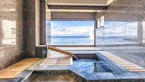 Un área de piscina interior moderna con vistas al mar, que incluye una terraza de madera, paredes de piedra y un recinto de vidrio.