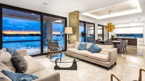 A modern living room with a large glass window, a beige sofa, a glass coffee table, and a dining area with a kitchen in the background. - Res & Campo (Pinhal Novo)