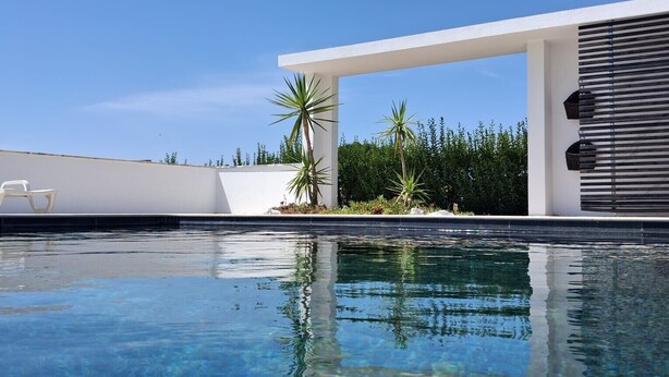 A clear blue pool with a modern white structure and palm trees in the background. - Res & Campo (Pinhal Novo)