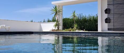 Piscina azul cristalina com uma estrutura branca moderna e palmeiras ao fundo.