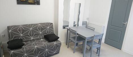 A small, modern room with a grey sofa, two black cushions, a dining table with four chairs, and a grey door.
