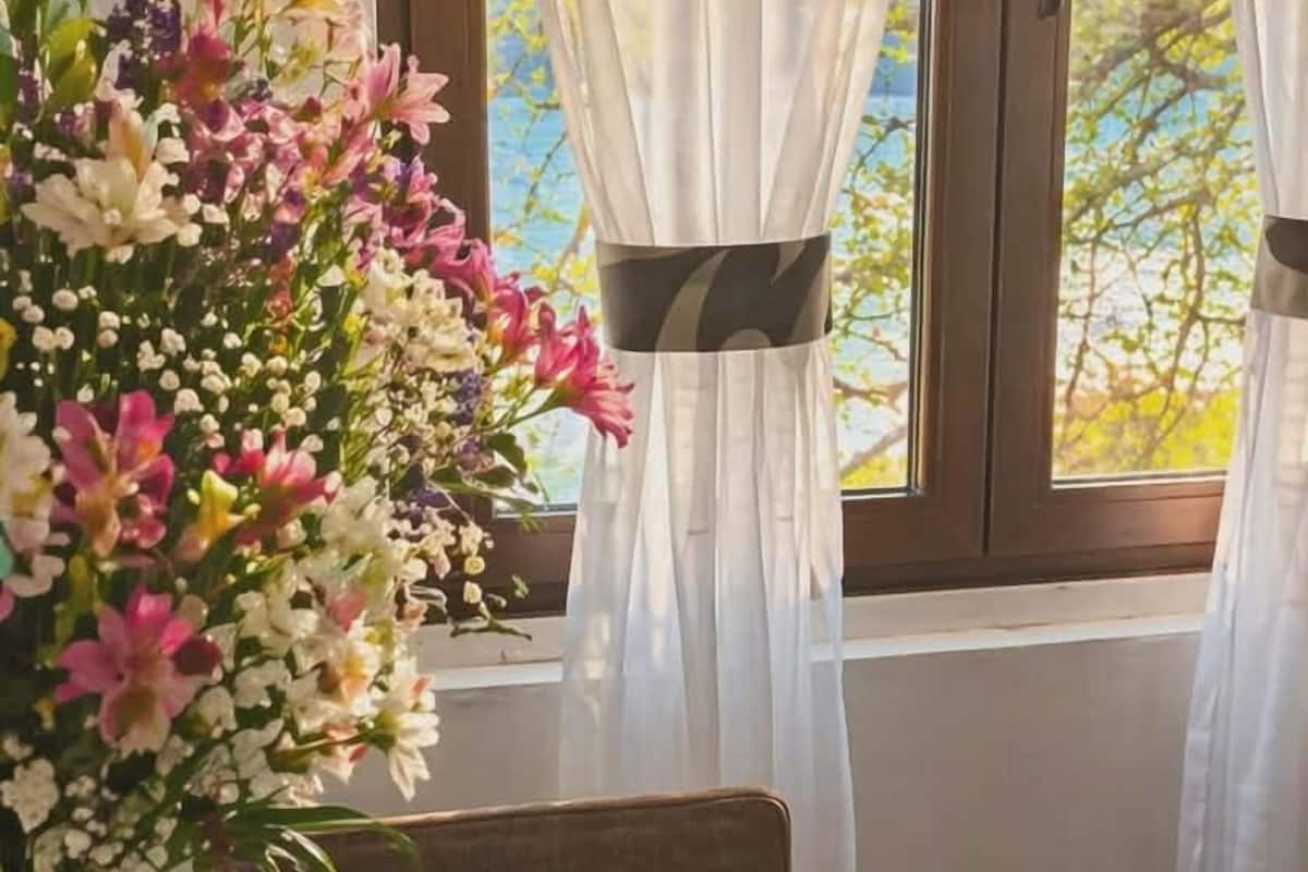 Una mesa con un arreglo floral, copas de vino y una botella, frente a una ventana con cortinas translúcidas y vista a árboles.