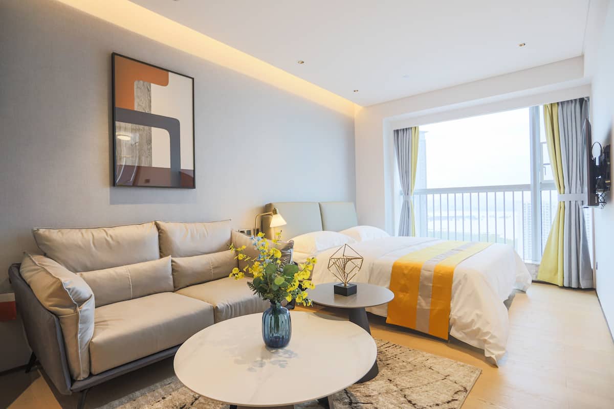 Una habitación de hotel moderna con un sofá, una mesa redonda de centro con un jarrón de flores, una cama con colcha amarilla y blanca, y un ventanal con cortinas.