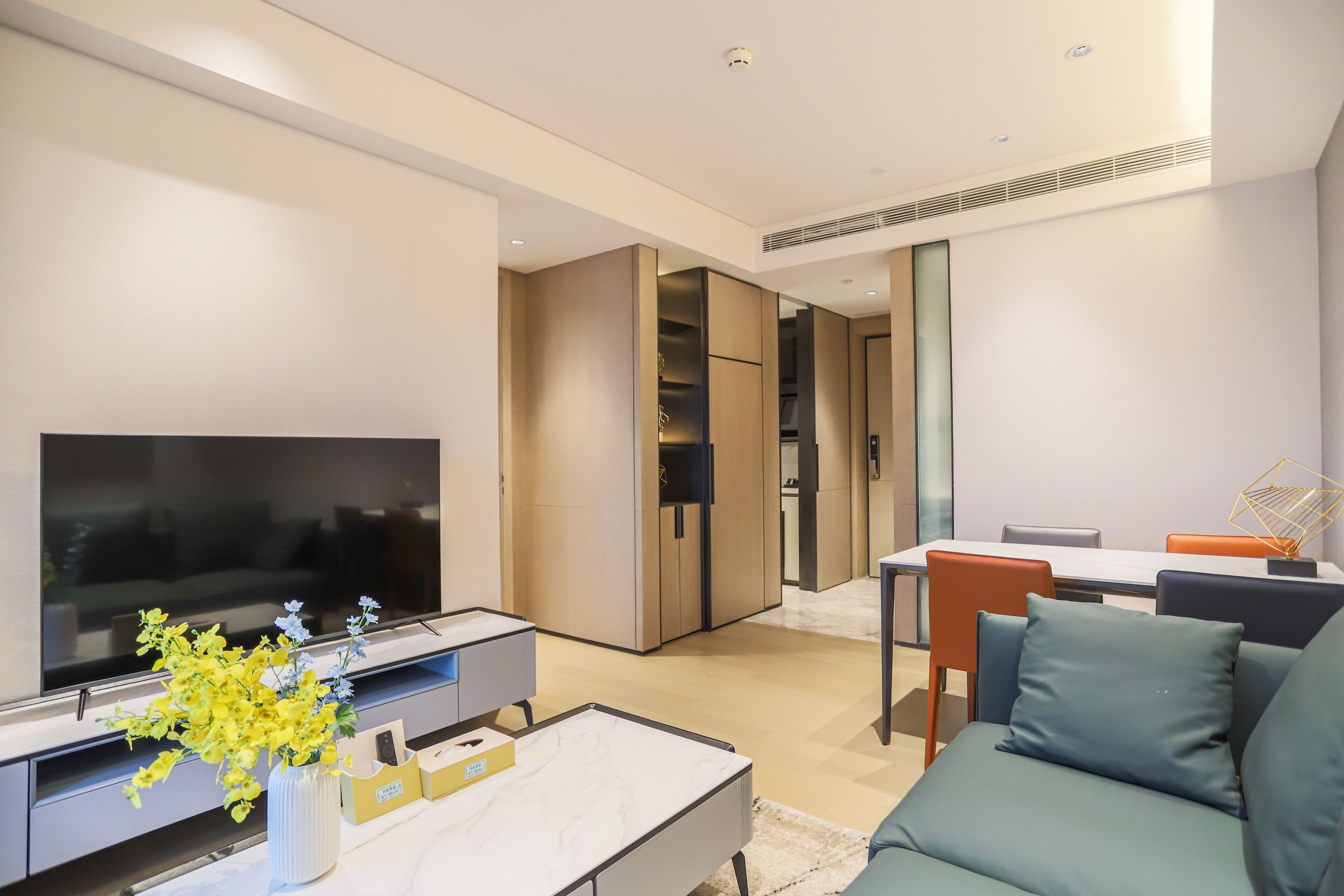 A modern living room with a flat-screen TV, a coffee table, and a sofa. There is a dining area with a table and chairs.