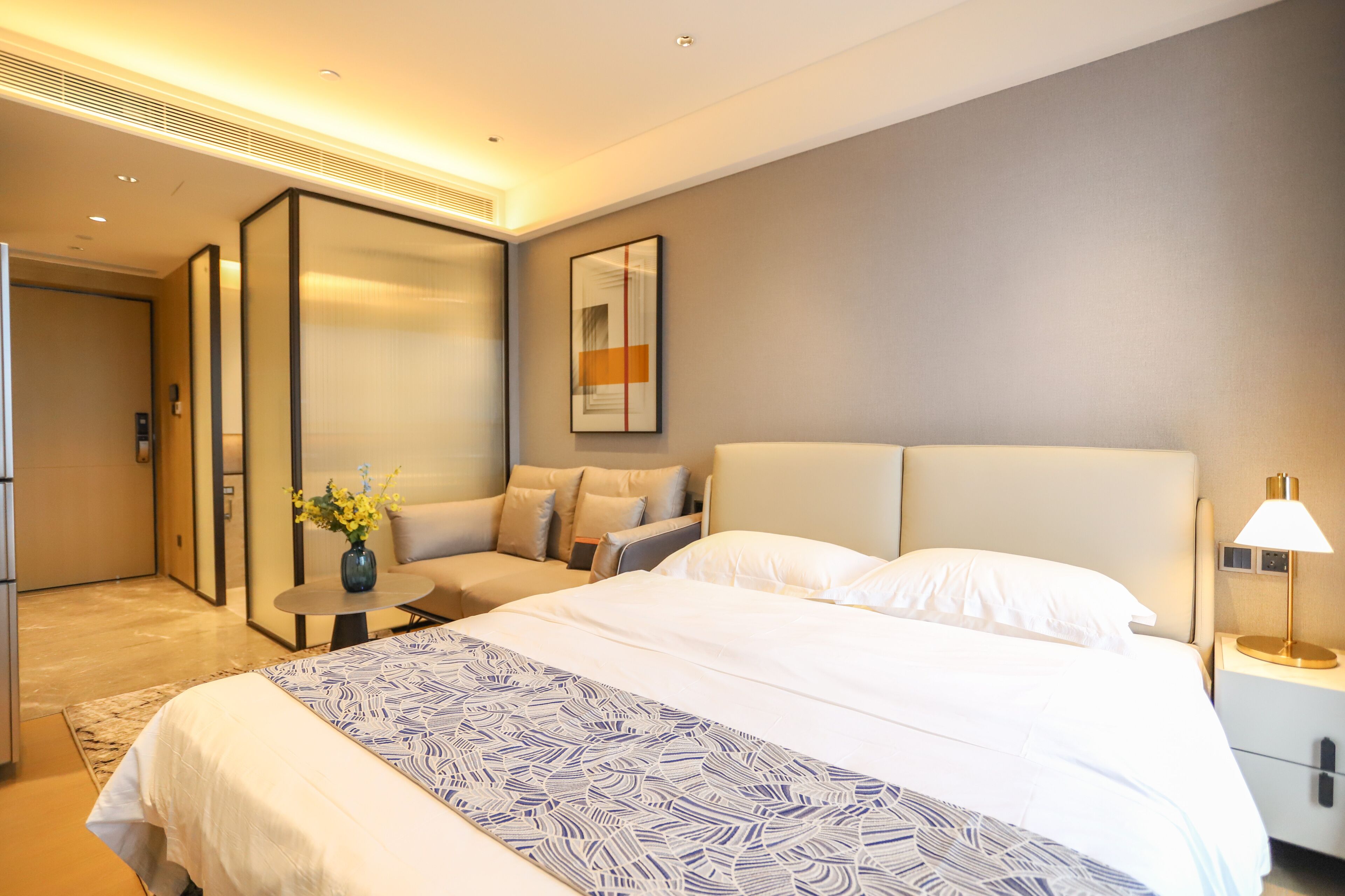 A modern hotel room with a large bed, a seating area, a small table with a vase of flowers, and a nightstand with a lamp.