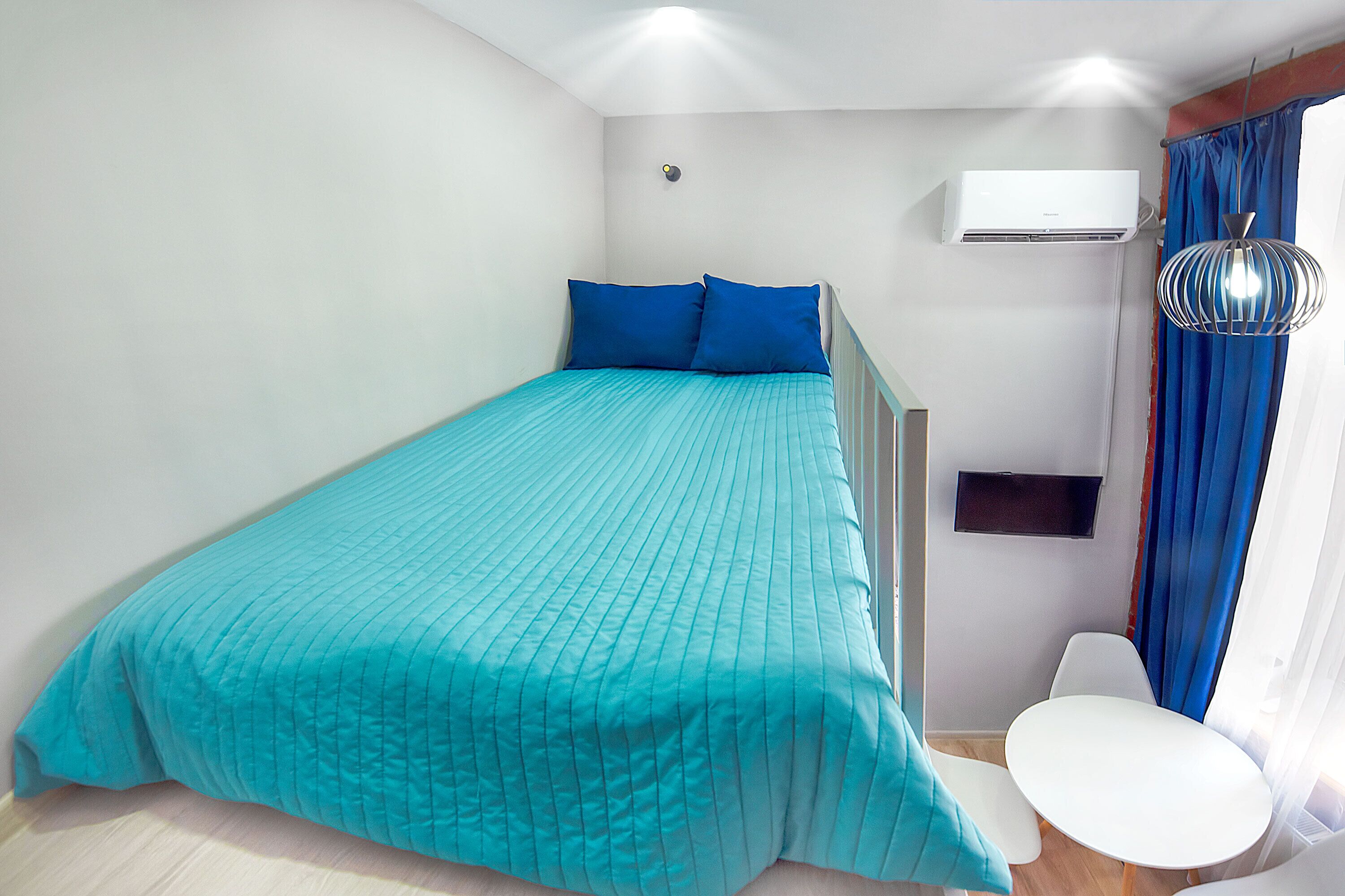 A bedroom with a bed, a white chair, a small table, an air conditioning unit, and a hanging light fixture.