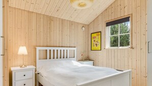 A bedroom with a wooden paneled wall, a bed with a white headboard, a bedside table with a lamp, a framed picture, and a window.