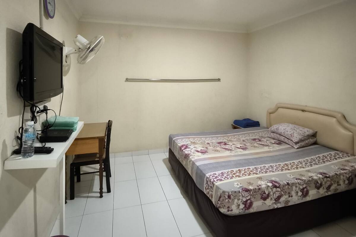 Une chambre à coucher avec un lit, un bureau avec un ordinateur, une chaise, une télévision et un ventilateur fixé au mur.