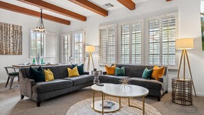 Ein Wohnzimmer mit Ecksofa, einem runden Couchtisch, einem Esstisch mit Stühlen und großen Fenstern mit Rollläden.